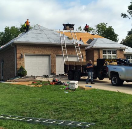 Workers on a roof under construction above a two-car garage, with ladders, a truck, and a trailer in the driveway.