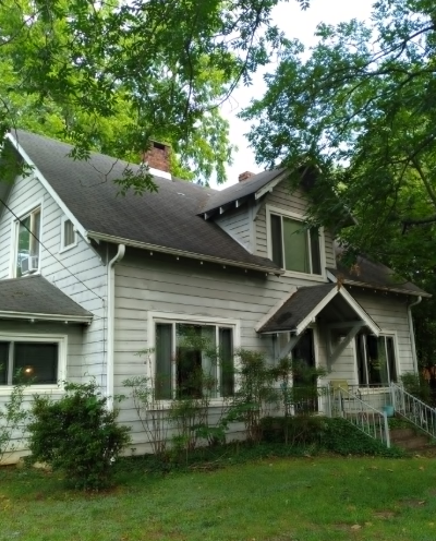 A light blue, two-story house with a grey roof, a chimney, and a small front porch, surrounded by green trees.