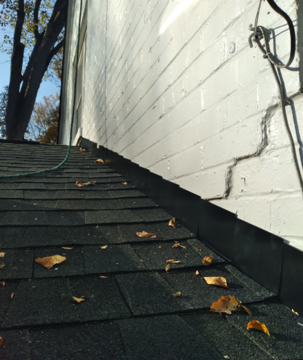 An angled view of a dark shingled roof meeting a white brick wall with a visible crack and black flashing.