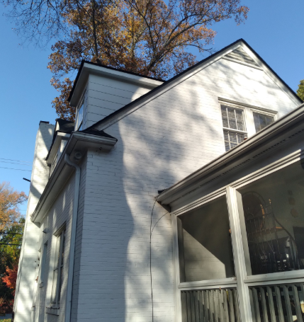 A low-angle view of a white brick, two-story house with a screened-in porch, framed against a clear blue sky and trees.