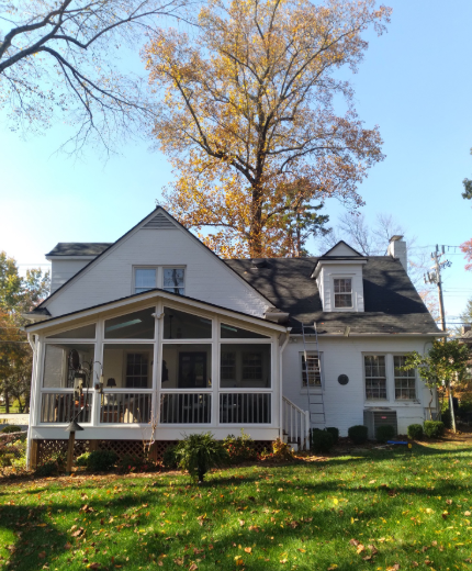A white house with a screened-in porch, a gabled roof, and a dormer window, set against a backdrop of autumn trees.