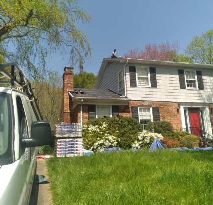 A construction worker on the roof of a two-story brick and siding house with a stack of shingles nearby and a work van.