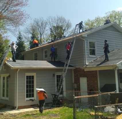 A crew of workers in bright safety shirts installing a new roof on a light gray house during a sunny day.