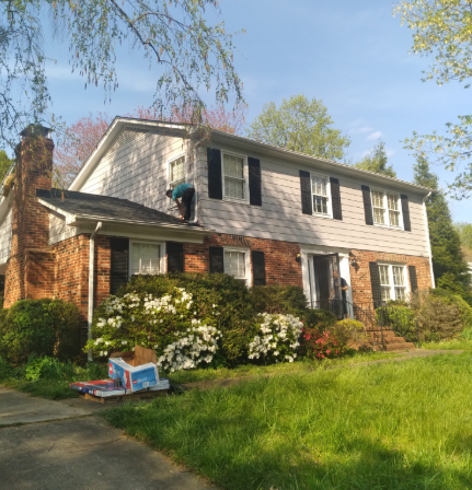 A person on a house roof near a second-story window, with a brick and light-sided exterior and white flowering bushes.
