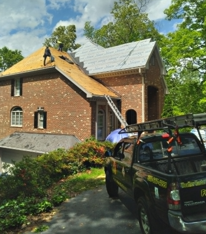 Workers replace the roof of a brick house while a company truck is parked in the driveway.