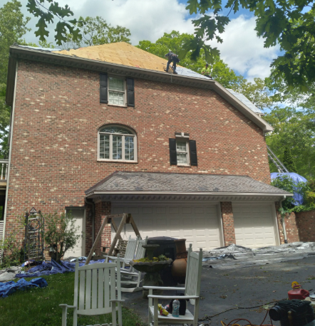 A two-story brick house with a partially replaced shingle roof where a worker is installing new wooden roof decking.