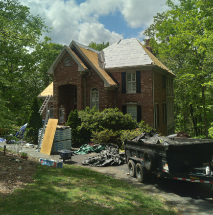 A brick house under renovation with a dumpster trailer in the driveway and roof materials scattered on the lawn.