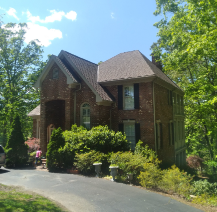 A two-story brick house with a steep shingled roof, surrounded by green trees, nestled in a landscaped yard.