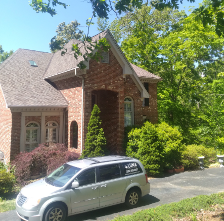 A silver minivan parked on a driveway in front of a multi-story brick house surrounded by trees.