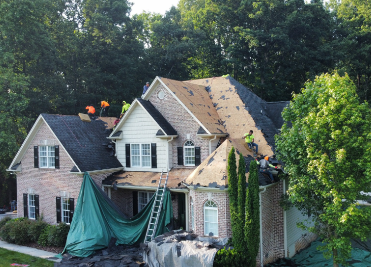 Construction workers in high-visibility vests replace shingles on the roof of a large brick house with green tarps below.