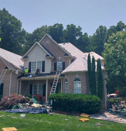 Workers replace the roof of a two-story brick house surrounded by trees on a sunny day.