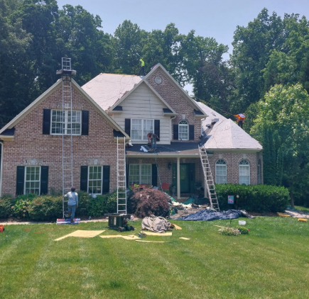 Workers perform roof repairs on a two-story brick house with ladders and equipment on the lawn.