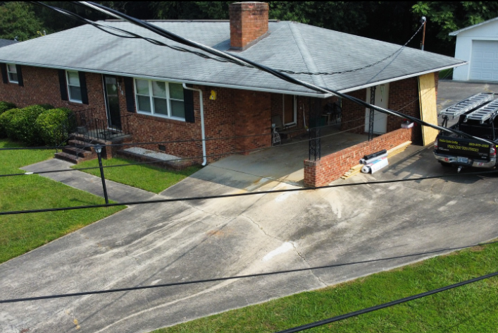 A red-brick ranch house with a carport and driveway, featuring a chimney and green lawn under a bright sky.