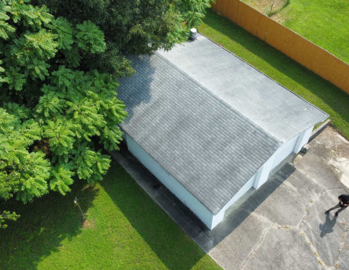 An aerial view of a rectangular building with a split roof, next to a large tree, grass, and a paved area.