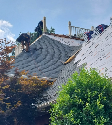 Two workers install gray asphalt shingles on a roof near a deck railing on a sunny day.