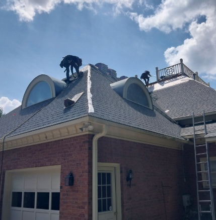 Two workers in safety gear repair the roof of a brick house with two dormer windows and a rooftop railing.