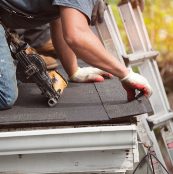A worker wearing gloves kneels on a roof, using a nail gun to install black shingles near a white gutter and ladder.