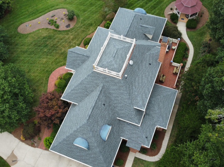 An aerial view of a large residential home with a grey shingled roof, a rectangular cupola, and surrounding landscaping.