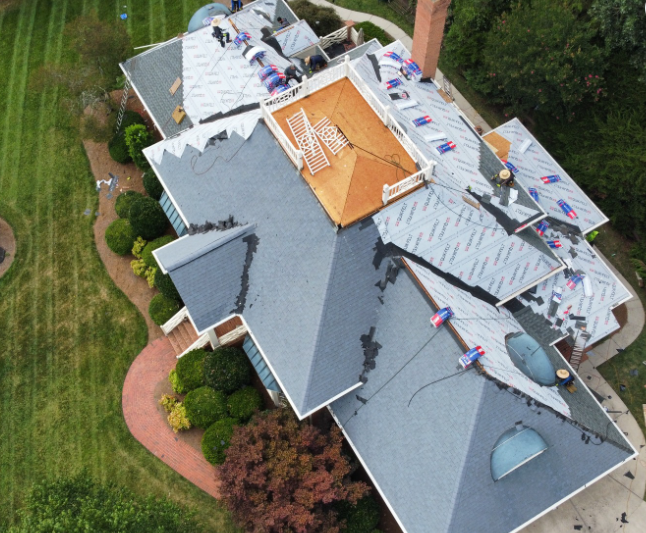 An aerial view shows a house undergoing roof repairs, with some sections covered in underlayment and others with shingles.