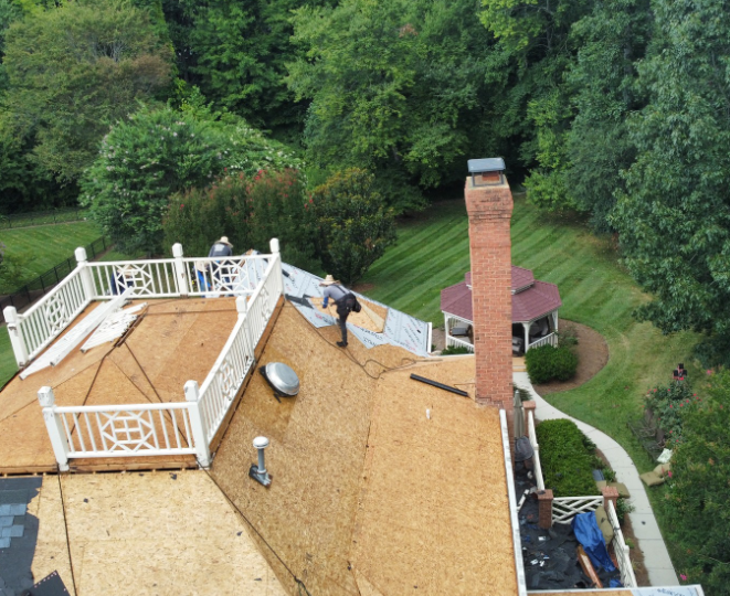 A high-angle view of a residential roof undergoing repair, featuring plywood decking, a white railing, and a brick chimney.