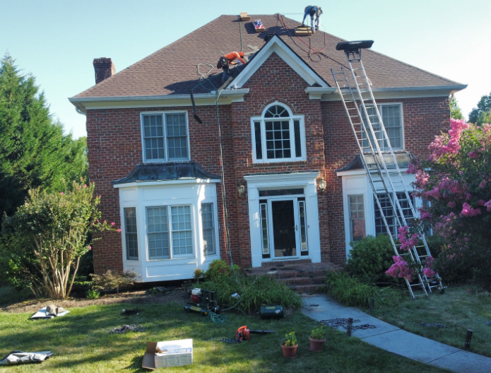 Roofers working on the roof of a two-story red brick house with a white bay window and a ladder leaning against the side.