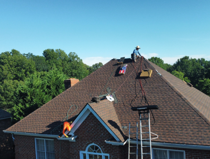 Two roofers work on a residential roof; one is near the peak with a harness, and the other is near a lower eave.