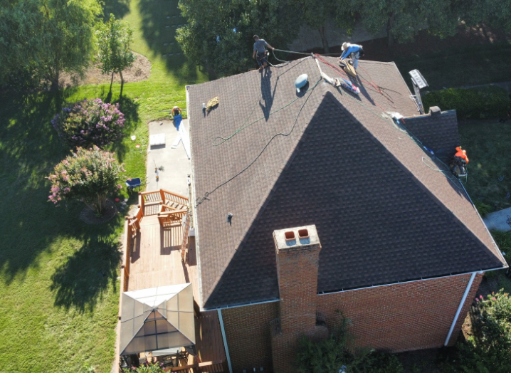 An aerial view of a brick house with workers repairing the dark shingled roof on a sunny day.