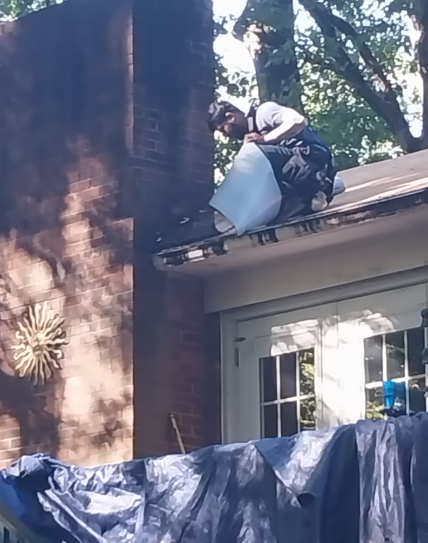 A person sits on a residential roof near a brick chimney, working with materials near the gutter line.