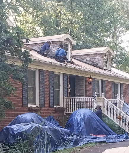 Two workers on a brick house roof installing shingles, with blue tarps covering the ground area below.