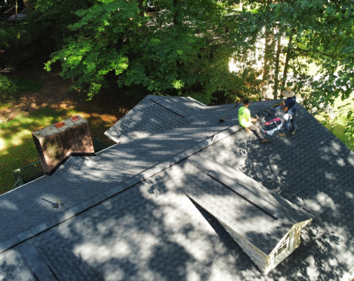 An aerial view shows two roofers working on a dark shingled roof next to a brick chimney and a dormer in a wooded area.