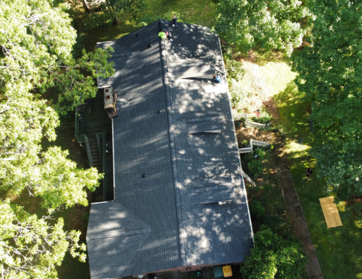 A top-down aerial view of a dark shingled residential roof surrounded by trees and a patch of grass.