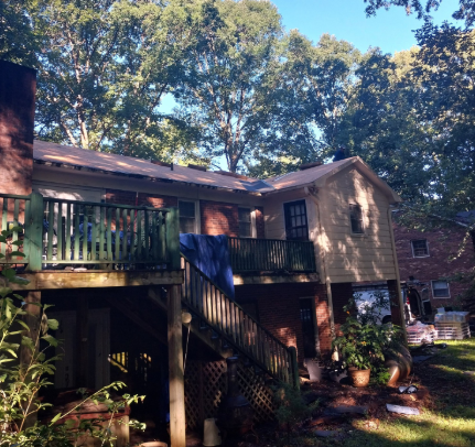 A two-story brick house features a wooden deck with green railings and a partially replaced, light-colored roof.