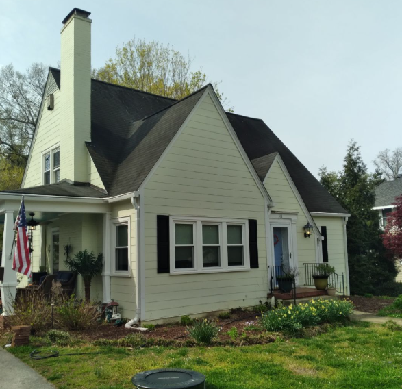 A pale yellow, two-story house with a prominent chimney, dark roof, and American flag on the front porch.