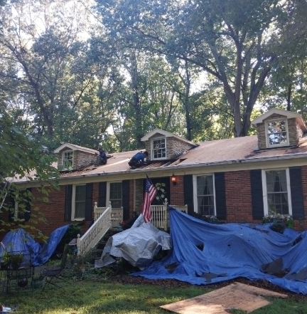 Roofers work on a brick house with dormer windows, surrounded by trees with blue tarps on the ground.