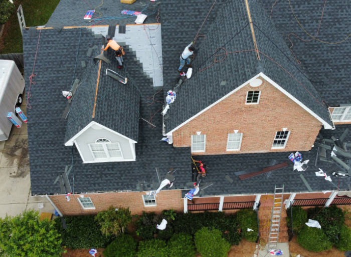 An aerial view shows workers installing dark shingles on a large residential roof with brick and siding sections.