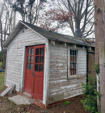 An old, white wooden shed with red double doors and a window, situated outdoors among trees on a cloudy day.
