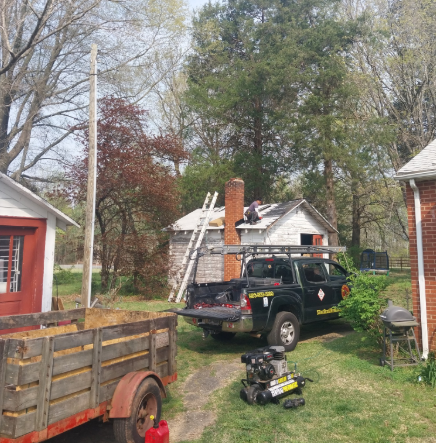 A worker repairs a residential roof next to a truck, a trailer, and trees in a grassy yard.