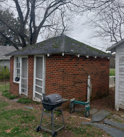 A small, single-story red brick outbuilding with a mossy, dark roof, screen doors, and a charcoal grill in the yard.