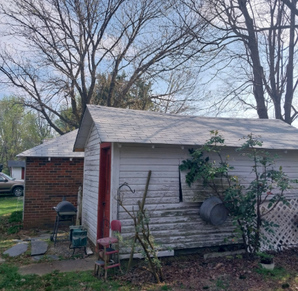 A weathered white wooden shed stands beside a brick building under a clear sky, with a small grill and chair nearby.