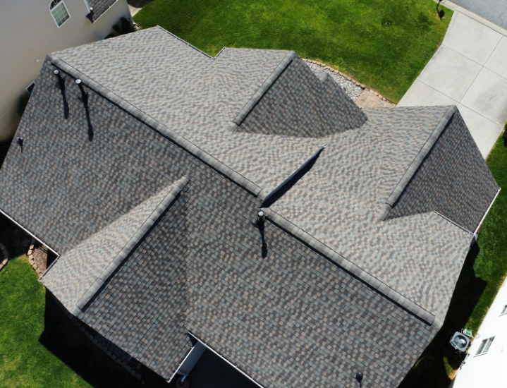 A high-angle aerial view of a gray shingled residential roof with multiple gables and vent pipes, surrounded by green lawns.