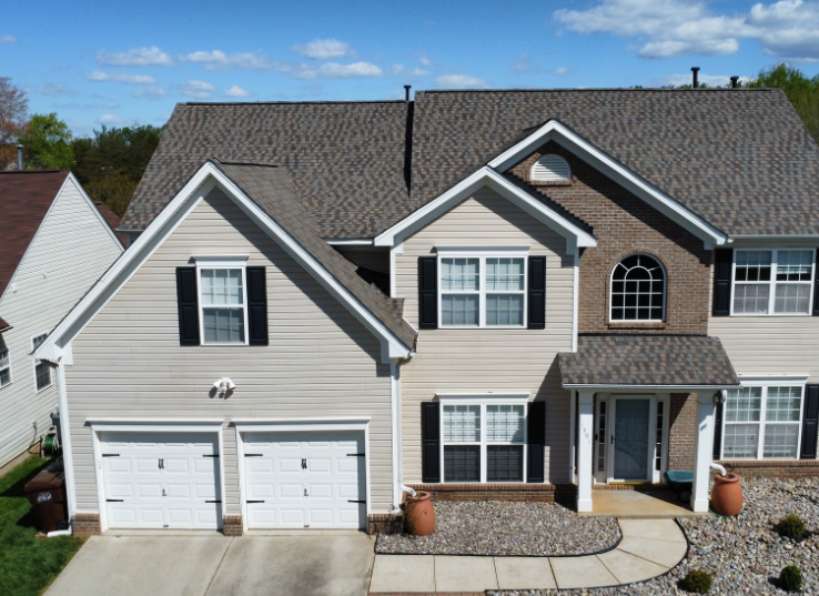 Two-story suburban house with beige siding, a brick facade, a two-car garage, and a grey shingled roof under a blue sky.