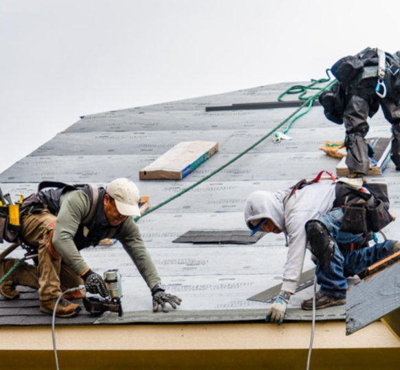 Three workers in protective gear install shingles on a sloped roof under a cloudy sky.