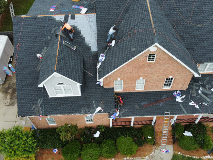 Aerial view of a crew installing black asphalt shingles on the roof of a large brick house.