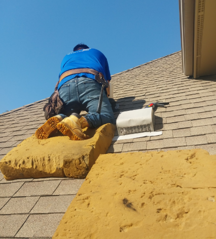 A worker wearing blue kneeling on foam pads on a shingled roof while repairing a vent.