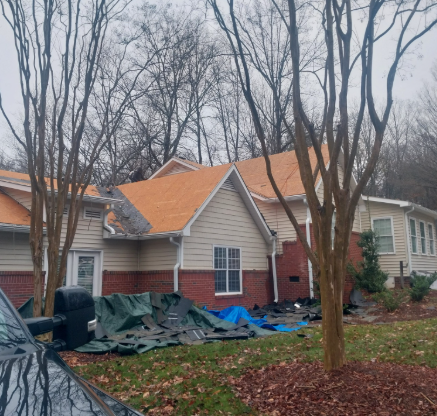 A residential house under roof renovation with orange underlayment exposed, surrounded by debris tarps and trees.
