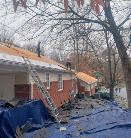 Two workers perform a roof replacement on a brick house, with a ladder leaning against the eaves and debris on blue tarps.