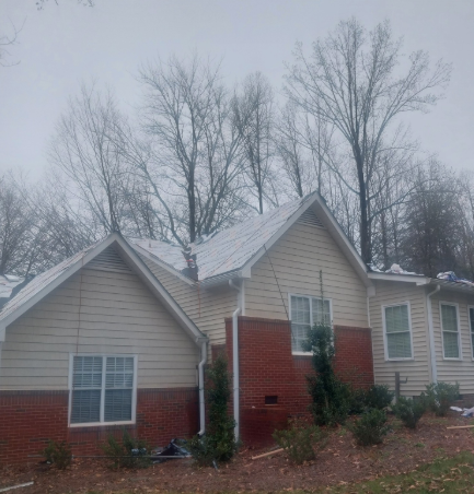 A one-story suburban home with tan siding and red brick, featuring gabled roofs against a background of bare trees.
