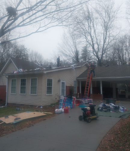 Workers on a roof and a ladder installing new shingles on a residential home with construction supplies on the driveway.