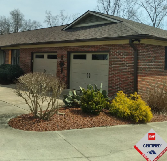 A brick garage with two light-colored doors and a brown shingled roof, fronted by a landscaped flower bed with shrubs.
