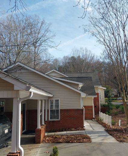 Exterior view of a one-story suburban home with tan siding, red brick, and a covered front entryway on a sunny day.